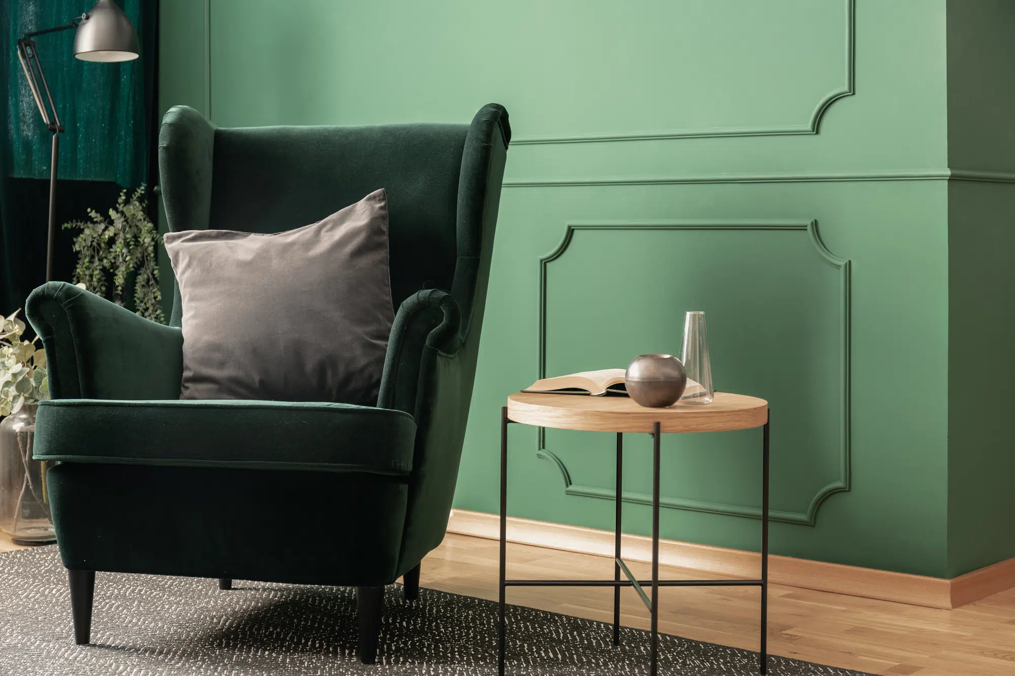 Close-up of a green velvet armchair with a gray cushion standing next to a wooden coffee table in a cozy living room interior with molding on the wall.