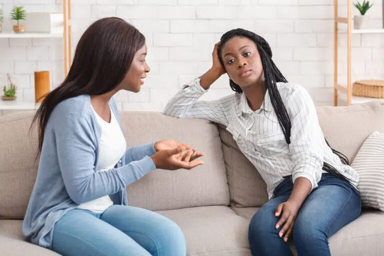 Bored black girl tired of listening her talkative girlfriend while sitting on sofa at home together